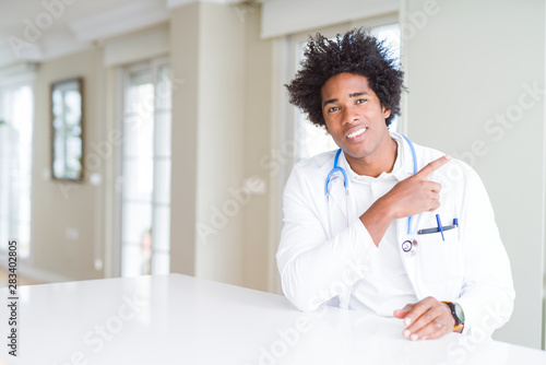 African American doctor man at the clinic cheerful with a smile of face pointing with hand and finger up to the side with happy and natural expression on face