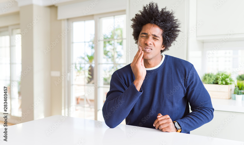 Young african american man wearing casual sweater sitting at home touching mouth with hand with painful expression because of toothache or dental illness on teeth. Dentist concept.