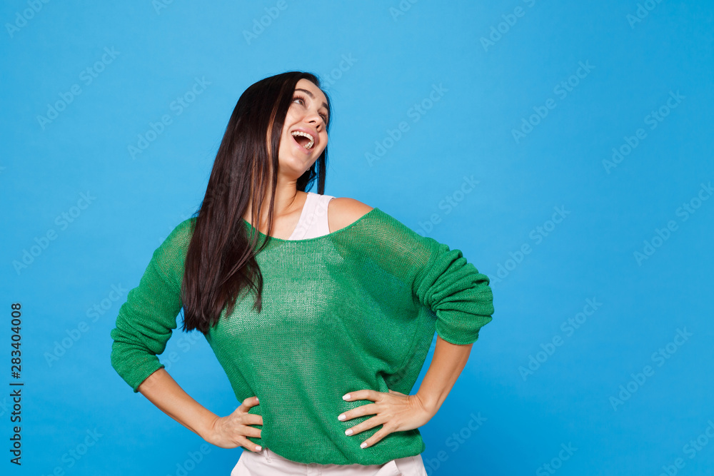 Φωτογραφία Portrait of cheerful young woman in green casual clothes looking up aside, standing with arms akimbo on waist isolated on blue wall background in studio