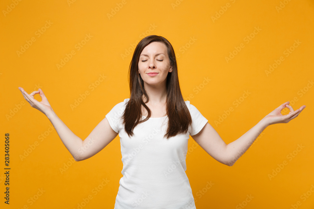 Relaxed young woman in white casual clothes keeping eyes closed hold hands in yoga gesture, relaxing meditating isolated on yellow orange wall background. People lifestyle concept. Mock up copy space. Relaxed young woman in white casual clothes keeping eyes closed hold hands in yoga gesture, relaxing meditating isolated on yellow orange wall background. People lifestyle concept. Mock up copy space.