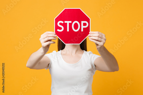 Portrait of young woman in white casual clothes covering face with red sign with Stop title in hands isolated on yellow orange wall background in studio. People lifestyle concept. Mock up copy space. photo