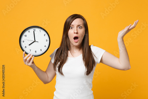 Portrait of shocked young woman in white casual clothes spreading hands, holding round clock isolated on bright yellow orange wall background in studio. People lifestyle concept. Mock up copy space.