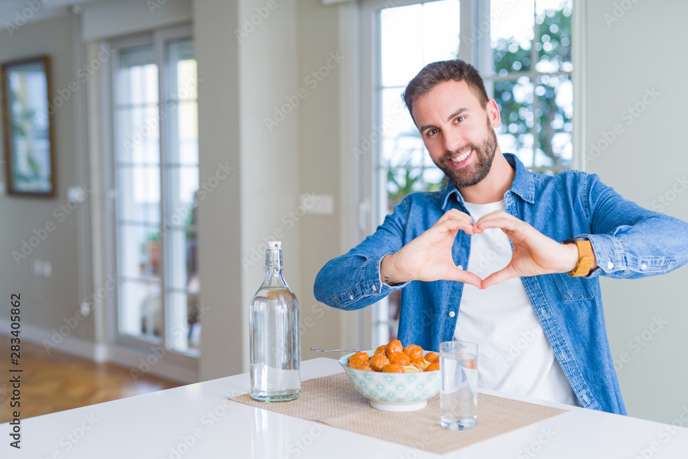 Handsome man eating pasta with meatballs and tomato sauce at home smiling in love showing heart symbol and shape with hands. Romantic concept. Handsome man eating pasta with meatballs and tomato sauce at home smiling in love showing heart symbol and shape with hands. Romantic concept.