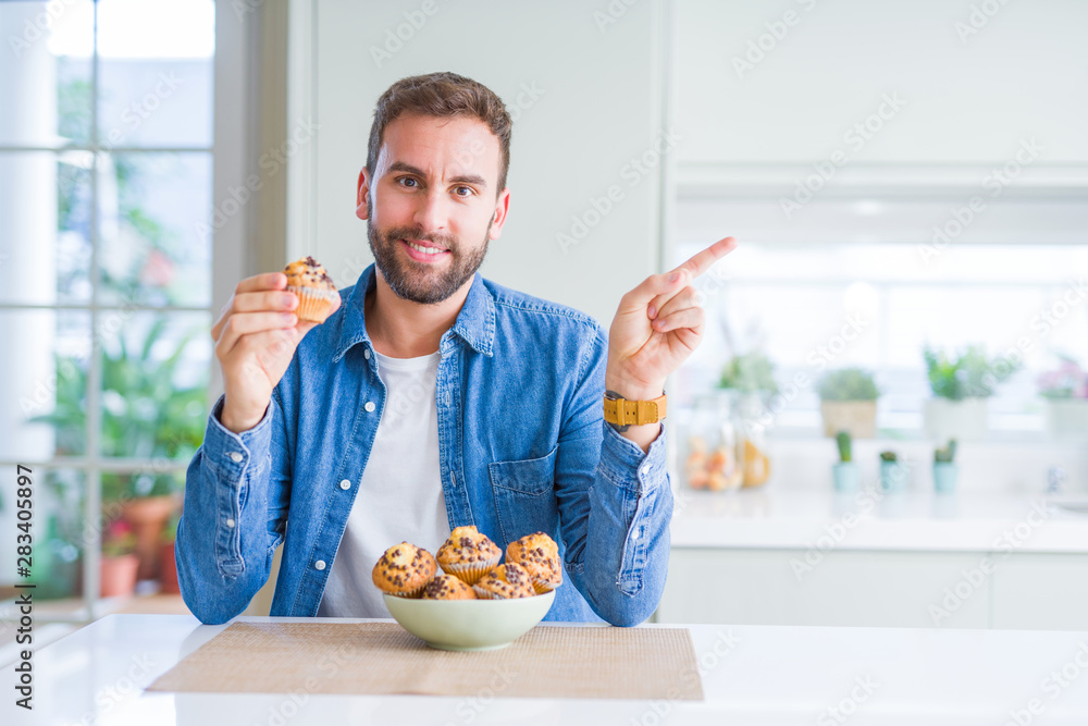 Handsome man eating chocolate chips muffin very happy pointing with hand and finger to the side Handsome man eating chocolate chips muffin very happy pointing with hand and finger to the side