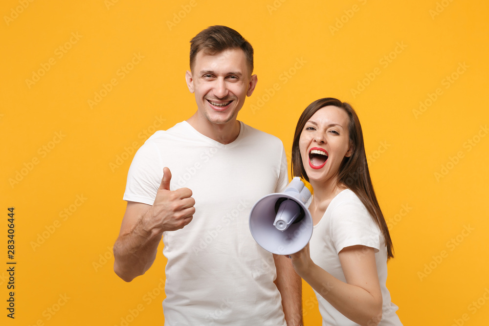 Cheerful young couple friends guy girl in white blank empty t-shirts posing isolated on yellow orange background. People lifestyle concept. Mock up copy space. Screaming in megaphone showing thumb up. Cheerful young couple friends guy girl in white blank empty t-shirts posing isolated on yellow orange background. People lifestyle concept. Mock up copy space. Screaming in megaphone showing thumb up.