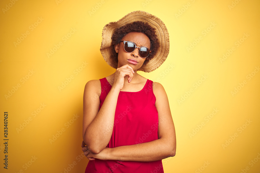 African american woman wearing summer hat and sunglasses over yellow isolated background with hand on chin thinking about question, pensive expression. Smiling with thoughtful face. Doubt concept. African american woman wearing summer hat and sunglasses over yellow isolated background with hand on chin thinking about question, pensive expression. Smiling with thoughtful face. Doubt concept.