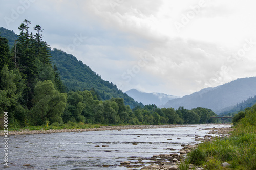 Mountain river with rapids and faults, slopes with coniferous trees, rocks sticking out of the water, the sky in the clouds