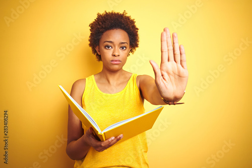 African american woman reading a book over yellow isolated background with open hand doing stop sign with serious and confident expression, defense gesture