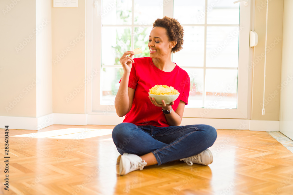 Young beautiful african american woman with afro hair eating chips sitting on the floor at home