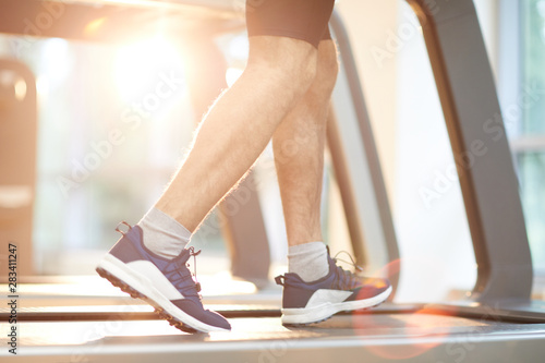Sports background of unrecognizable man running on treadmill in gym lit by sunlight, copy space