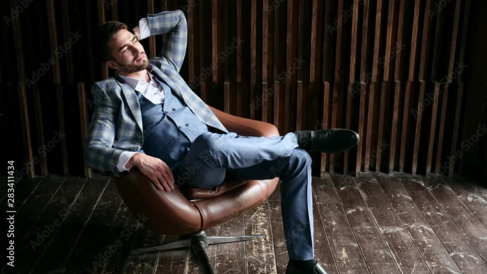 Young handsome businessman with a beard in a chic blue suit is resting. the lawyer is sitting in a leather chair by the window. Young handsome businessman with a beard in a chic blue suit is resting. the lawyer is sitting in a leather chair by the window.