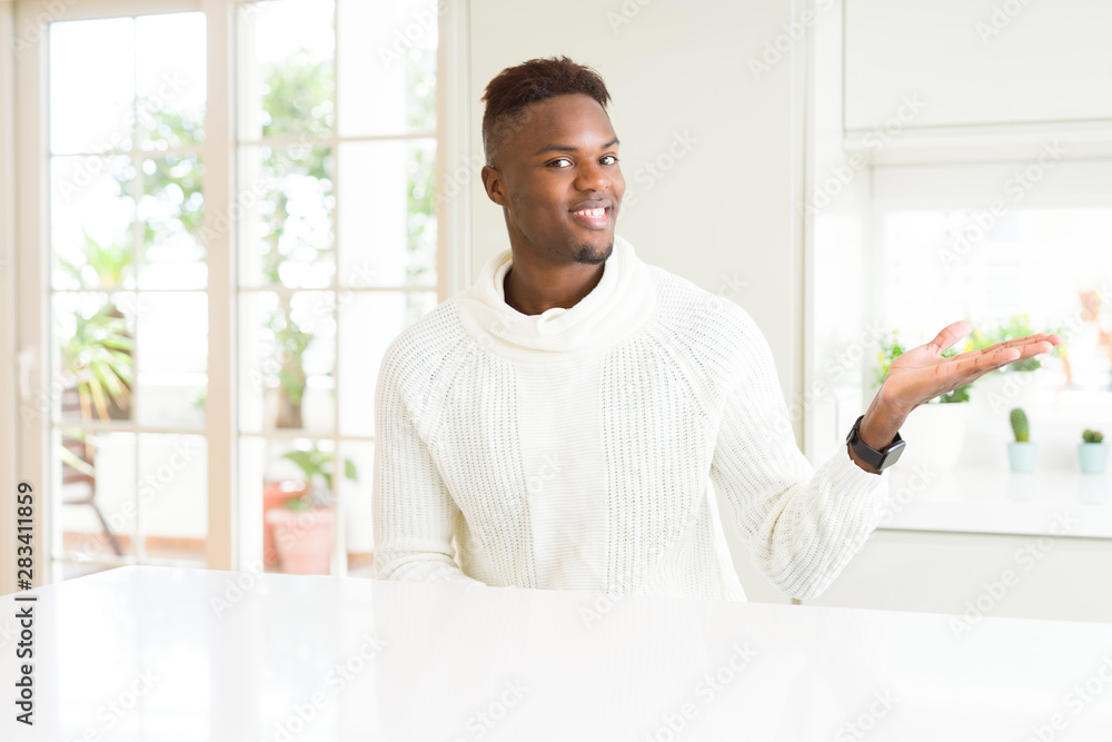 Handsome african american man on white table smiling cheerful presenting and pointing with palm of hand looking at the camera. Handsome african american man on white table smiling cheerful presenting and pointing with palm of hand looking at the camera.