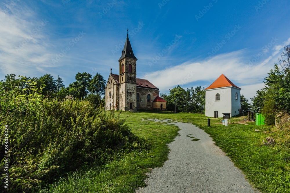 Krasikov, Kokasice / Czech Republic - August 9 2019: View of the church of Mary Magdalene and a bell tower. Bright sunny day with blue sky and white clouds. Gravel footh path leading to church.