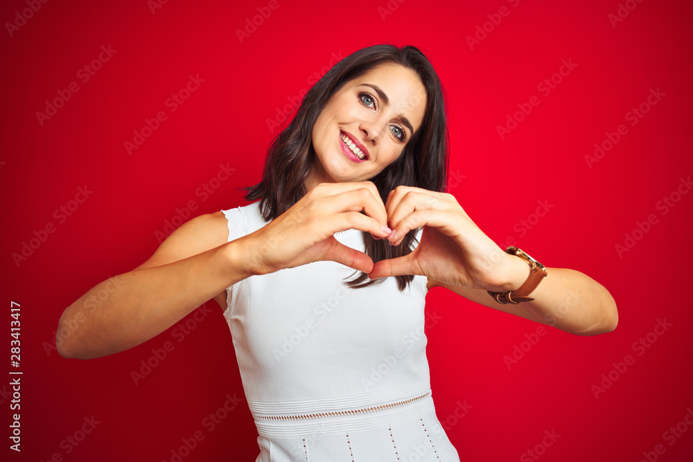 Young beautiful woman wearing white dress standing over red isolated background smiling in love doing heart symbol shape with hands. Romantic concept. Young beautiful woman wearing white dress standing over red isolated background smiling in love doing heart symbol shape with hands. Romantic concept.