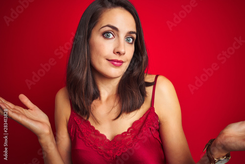 Young beautiful woman wearing sexy lingerie over red isolated background clueless and confused expression with arms and hands raised. Doubt concept.