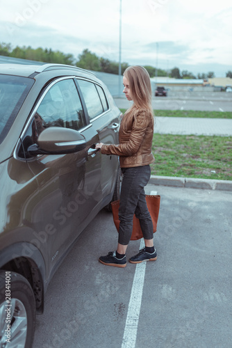 girl in a jacket and jeans opens car door in parking lot near a shopping center. Alarm activation. SUV crossover green in summer in city. Return from store with bag and shopping. © byswat