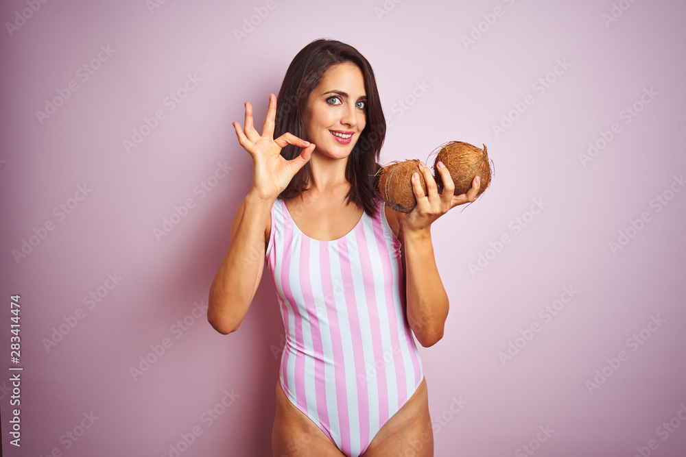 Beautiful woman wearing pink striped swimsuit holding coconuts over isolated pink background doing ok sign with fingers, excellent symbol Beautiful woman wearing pink striped swimsuit holding coconuts over isolated pink background doing ok sign with fingers, excellent symbol