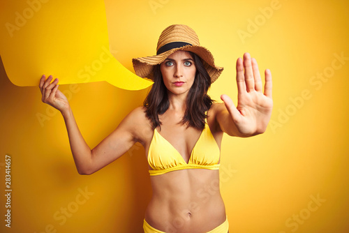Beautiful woman wearing yellow bikini and holding talking balloon over isolated yellow background with open hand doing stop sign with serious and confident expression, defense gesture © Krakenimages.com