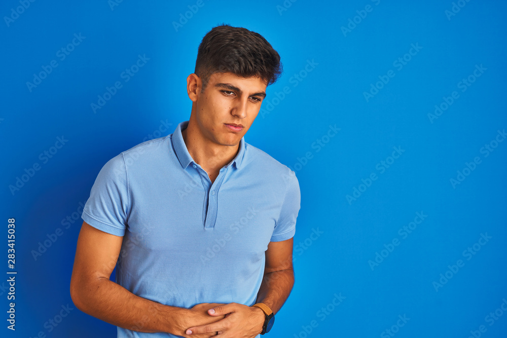 Young indian man wearing casual polo standing over isolated blue background with hand on stomach because nausea, painful disease feeling unwell. Ache concept. Young indian man wearing casual polo standing over isolated blue background with hand on stomach because nausea, painful disease feeling unwell. Ache concept.