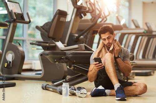 Full length portrait of handsome man sitting on floor in gym and using smartphone, copy space