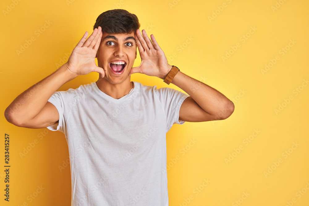 Young indian man wearing white t-shirt standing over isolated yellow background Smiling cheerful playing peek a boo with hands showing face. Surprised and exited Young indian man wearing white t-shirt standing over isolated yellow background Smiling cheerful playing peek a boo with hands showing face. Surprised and exited