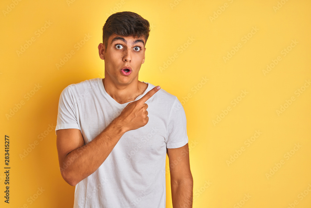 Young indian man wearing white t-shirt standing over isolated yellow background Surprised pointing with finger to the side, open mouth amazed expression. Young indian man wearing white t-shirt standing over isolated yellow background Surprised pointing with finger to the side, open mouth amazed expression.