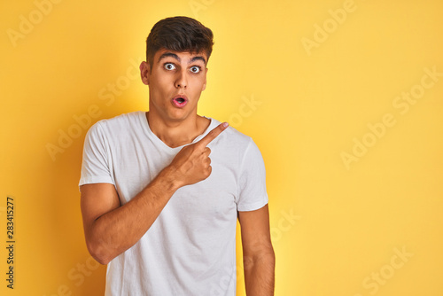 Young indian man wearing white t-shirt standing over isolated yellow background Surprised pointing with finger to the side, open mouth amazed expression.