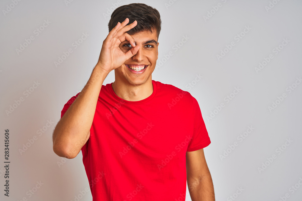 Young indian man wearing red t-shirt standing over isolated white background doing ok gesture with hand smiling, eye looking through fingers with happy face. Young indian man wearing red t-shirt standing over isolated white background doing ok gesture with hand smiling, eye looking through fingers with happy face.