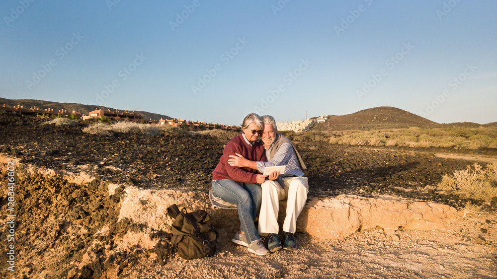Fototapeta premium Attractive couple of senior people sitting together on the cliff in front to the sea. Looking at tablet and smiling. Sunrise and arid landscape as background