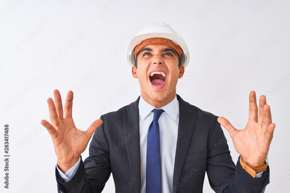 Young handsome architect man wearing suit and helmet over isolated white background crazy and mad shouting and yelling with aggressive expression and arms raised. Frustration concept.