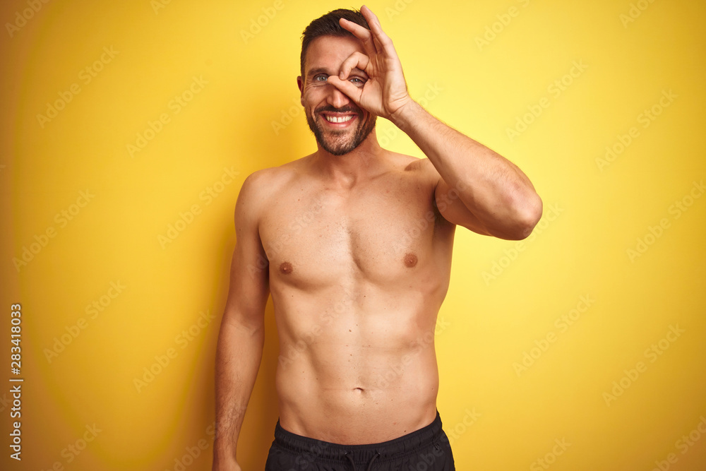 Young handsome shirtless man over isolated yellow background doing ok gesture with hand smiling, eye looking through fingers with happy face.