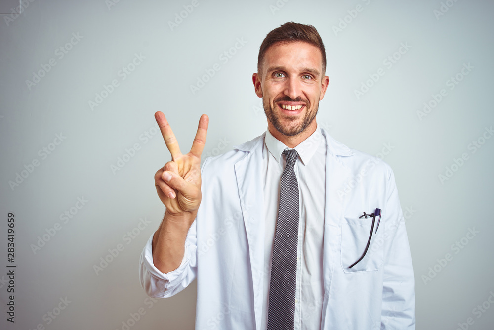 Young handsome doctor man wearing white profressional coat over isolated background showing and pointing up with fingers number two while smiling confident and happy. Young handsome doctor man wearing white profressional coat over isolated background showing and pointing up with fingers number two while smiling confident and happy.