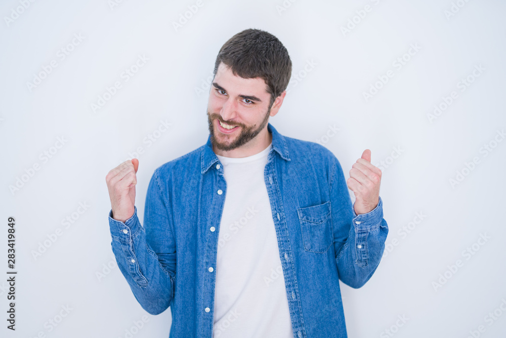 Young handsome hispanic man wearing denim jacket over white isolated background very happy and excited doing winner gesture with arms raised, smiling and screaming for success. Celebration concept. Young handsome hispanic man wearing denim jacket over white isolated background very happy and excited doing winner gesture with arms raised, smiling and screaming for success. Celebration concept.