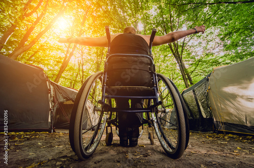 Disabled man resting in a campsite with friends. Wheelchair in the forest on the background of tents