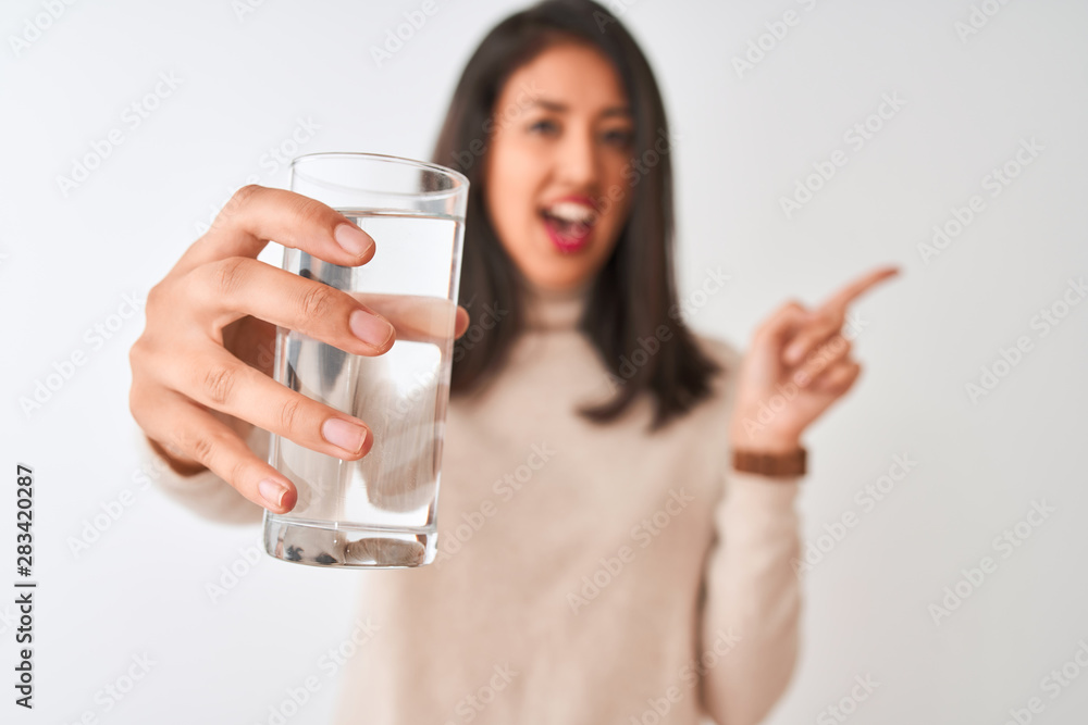 Young beautiful chinese woman holding glass of water standing over isolated white background very happy pointing with hand and finger to the side Young beautiful chinese woman holding glass of water standing over isolated white background very happy pointing with hand and finger to the side