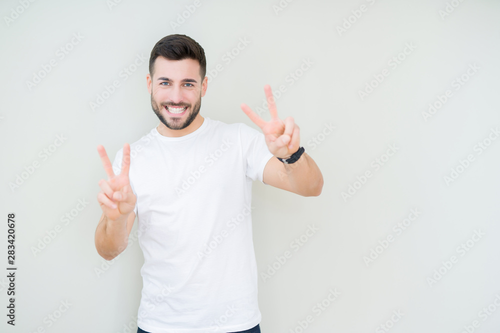 Young handsome man wearing casual white t-shirt over isolated background smiling looking to the camera showing fingers doing victory sign. Number two. Young handsome man wearing casual white t-shirt over isolated background smiling looking to the camera showing fingers doing victory sign. Number two.