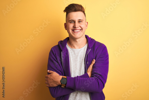 Young handsome man wearing purple sweatshirt standing over isolated yellow background happy face smiling with crossed arms looking at the camera. Positive person. © Krakenimages.com