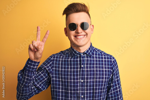 Young handsome man wearing casual shirt and sunglasses over isolated yellow background smiling with happy face winking at the camera doing victory sign. Number two.
