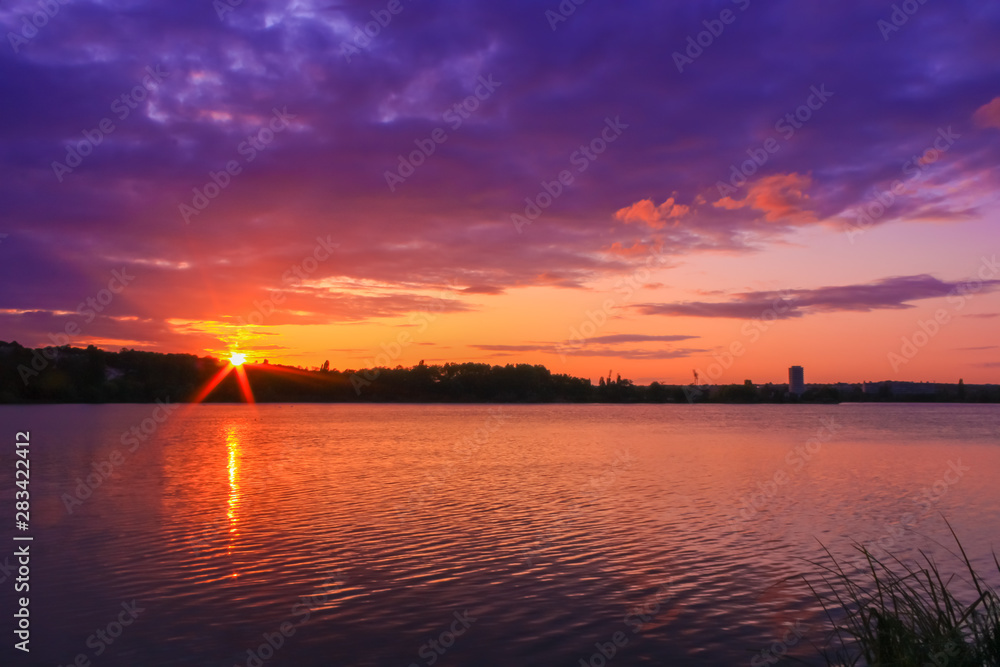 Nice colorful sunset over the water. Romantic landscape in front of a lake. Small ripples of water with reflection of sun rays. Vegetations on the side. 