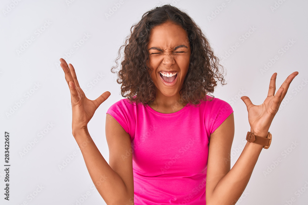Young brazilian woman wearing pink t-shirt standing over isolated white background celebrating mad and crazy for success with arms raised and closed eyes screaming excited. Winner concept Young brazilian woman wearing pink t-shirt standing over isolated white background celebrating mad and crazy for success with arms raised and closed eyes screaming excited. Winner concept