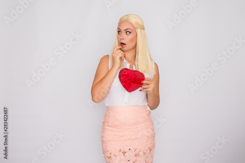 Portrait of a young blonde woman in light-colored clothes. Holding a heart shaped gift box.