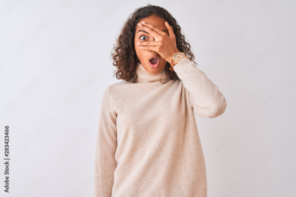 Young brazilian woman wearing turtleneck sweater standing over isolated white background peeking in shock covering face and eyes with hand, looking through fingers with embarrassed expression. Young brazilian woman wearing turtleneck sweater standing over isolated white background peeking in shock covering face and eyes with hand, looking through fingers with embarrassed expression.