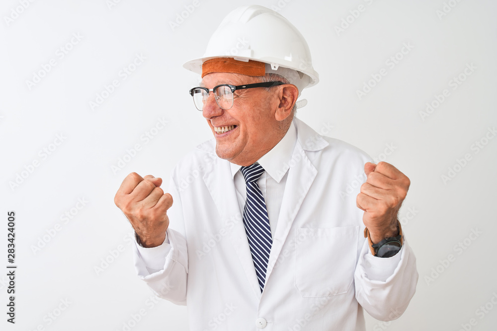 Senior grey-haired engineer man wearing coat and helmet over isolated white background very happy and excited doing winner gesture with arms raised, smiling and screaming for success. . Senior grey-haired engineer man wearing coat and helmet over isolated white background very happy and excited doing winner gesture with arms raised, smiling and screaming for success. .