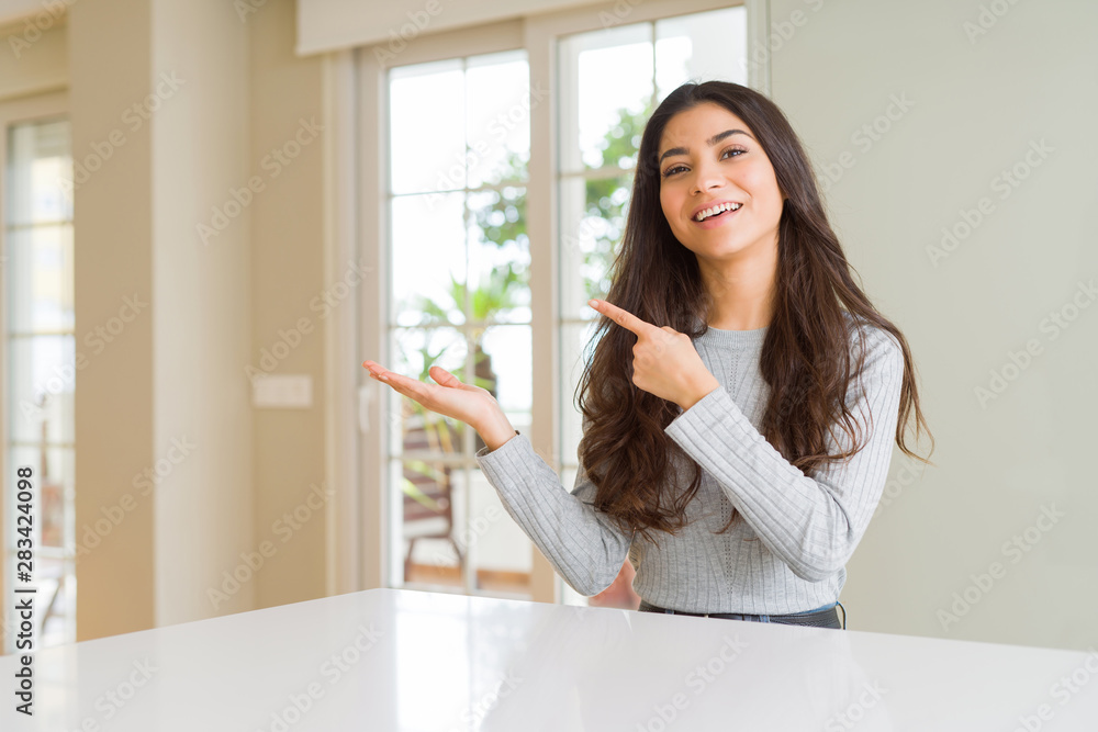 Young beautiful woman at home amazed and smiling to the camera while presenting with hand and pointing with finger. Young beautiful woman at home amazed and smiling to the camera while presenting with hand and pointing with finger.