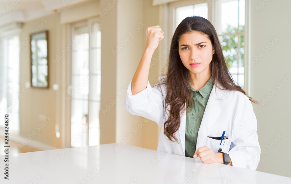 Young woman wearing medical coat at the clinic as therapist or doctor angry and mad raising fist frustrated and furious while shouting with anger. Rage and aggressive concept. Young woman wearing medical coat at the clinic as therapist or doctor angry and mad raising fist frustrated and furious while shouting with anger. Rage and aggressive concept.