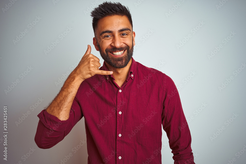Fototapeta premium Young indian man wearing red elegant shirt standing over isolated grey background smiling doing phone gesture with hand and fingers like talking on the telephone. Communicating concepts.