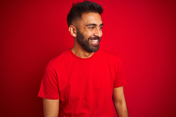 Young handsome indian man wearing t-shirt over isolated red background looking away to side with smile on face, natural expression. Laughing confident.