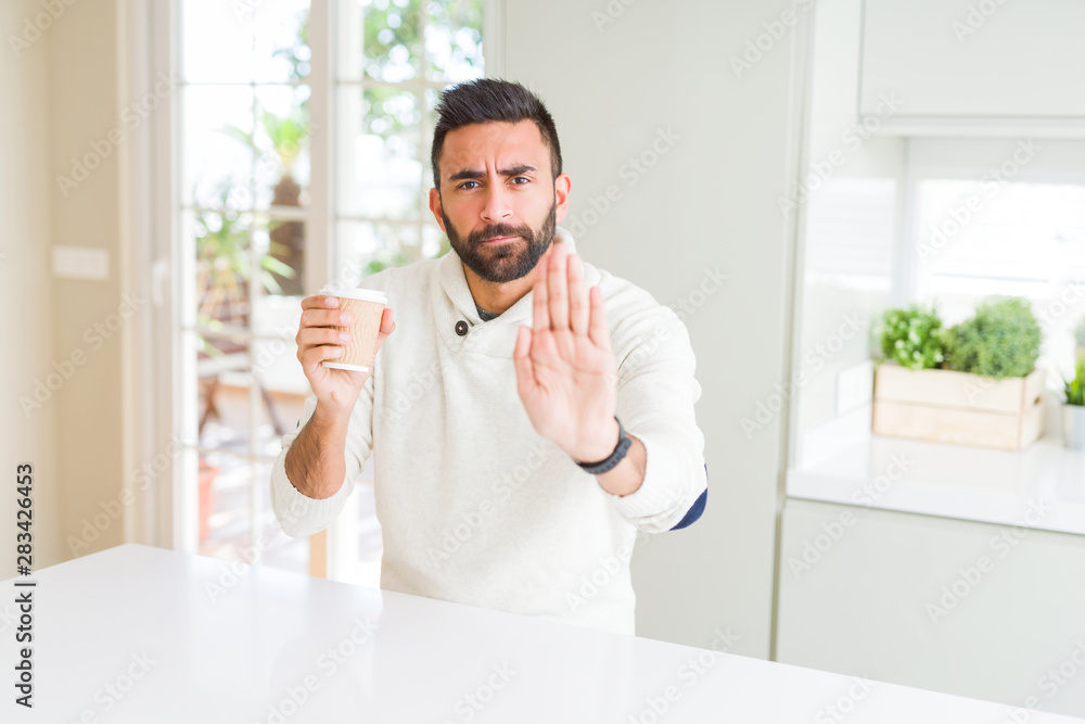 Handsome hispanic man drinking a coffee in a paper cup with open hand doing stop sign with serious and confident expression, defense gesture