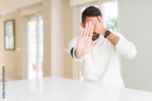 Handsome hispanic man wearing casual white sweater at home covering eyes with hands and doing stop gesture with sad and fear expression. Embarrassed and negative concept.