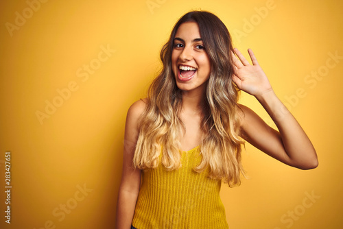 Young beautiful woman wearing t-shirt over yellow isolated background smiling with hand over ear listening an hearing to rumor or gossip. Deafness concept.